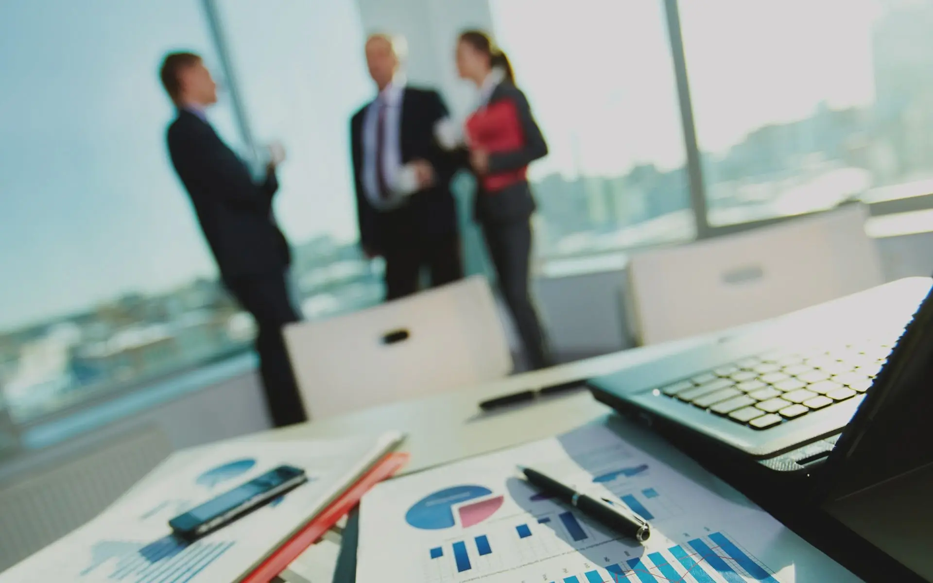 The image shows a business meeting in an office setting. A laptop, graphs, and a pen are in the foreground, while three blurred figures are in the background.